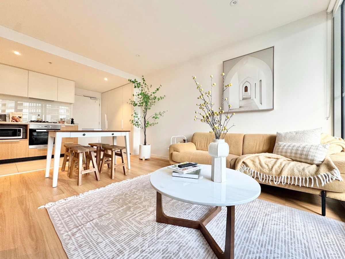 A stylish living area features a beige sofa adorned with a textured throw. A round coffee table sits centrally, surrounded by simple wooden stools. A bright, airy kitchen is visible in the background, complemented by natural light from large windows.