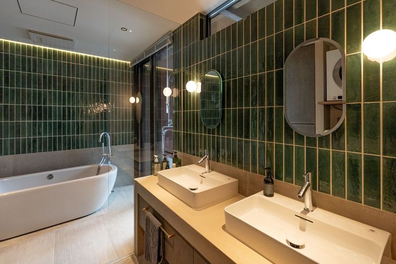 A bathroom with a serene aesthetic features two rectangular sinks situated on a light-colored countertop. A freestanding bathtub is positioned against a lush green tiled wall. Soft round lighting highlights the space, creating a calming ambiance.