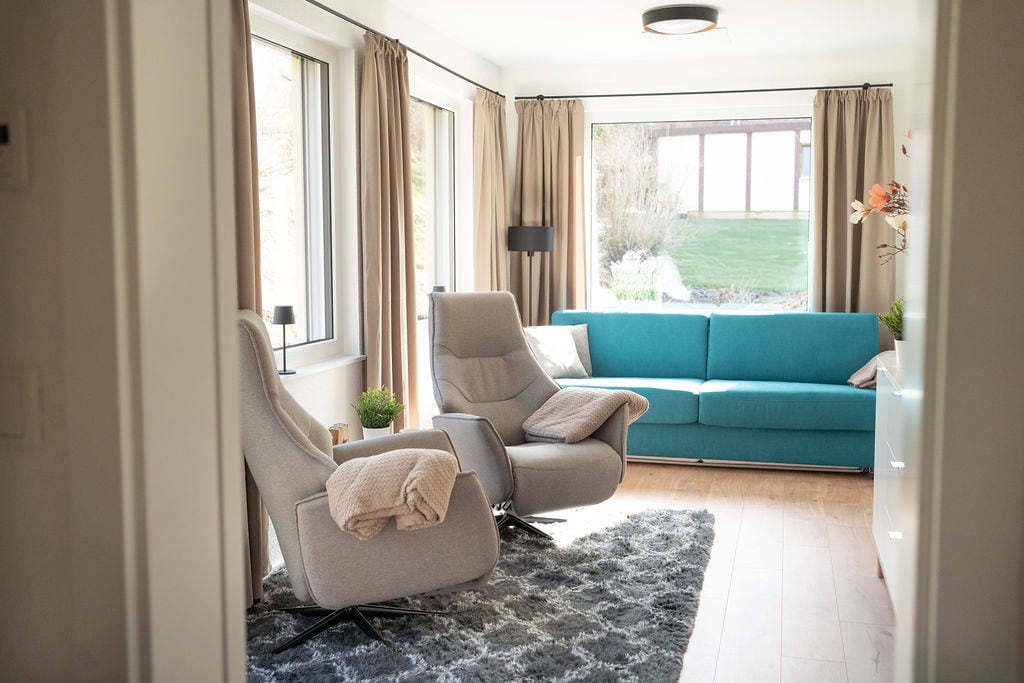 A modern living area features two comfortable lounge chairs and a vibrant blue sofa. Natural light enters through large windows, complemented by soft, beige curtains. A textured area rug is placed on the wooden floor, enhancing the open and inviting feel of the space.
