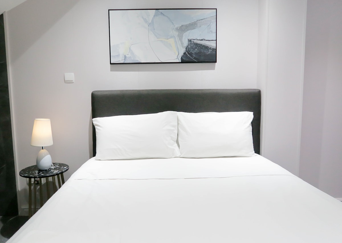 The room features a comfortable queen-sized bed dressed in crisp white linens. A modern artwork is displayed above the bed, complemented by a small side table with a lamp, creating a calming atmosphere in the cozy space.
