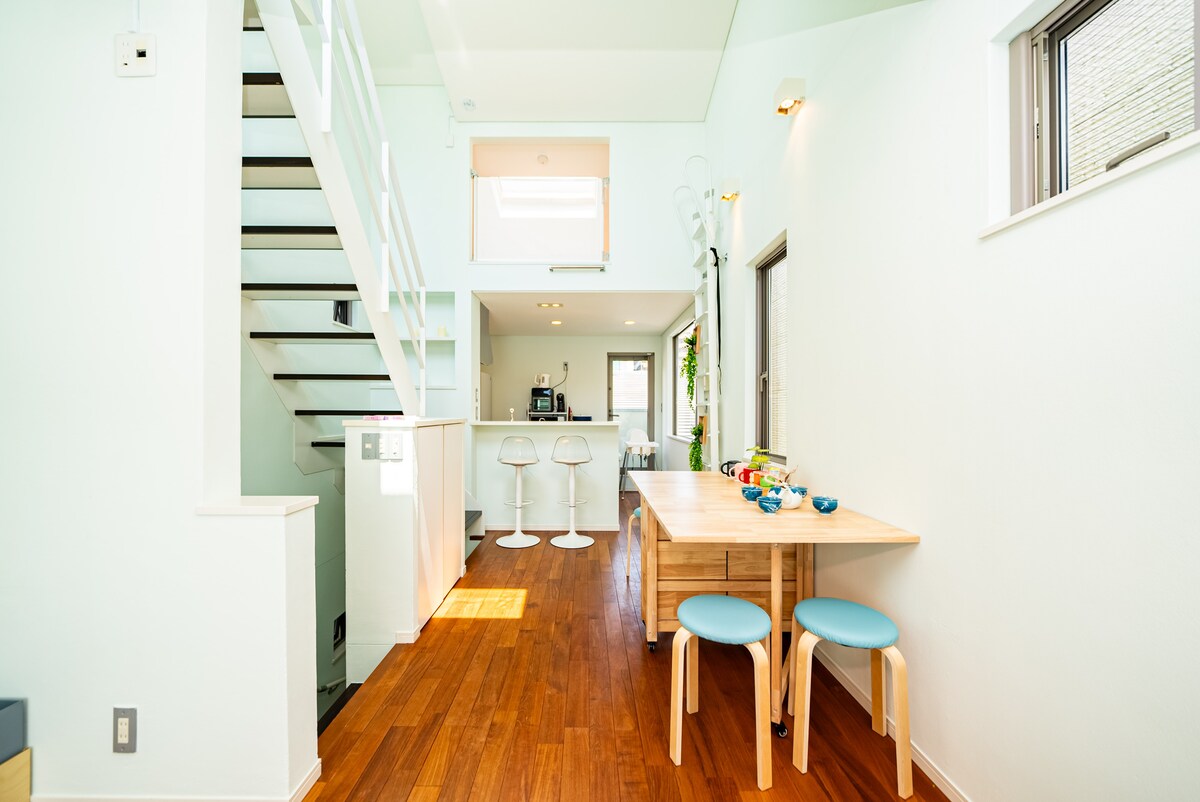 A bright and spacious dining area features a light wooden table with colorful dishes arranged on it. Two stools with light blue seats complement the natural wood. A modern kitchen is visible in the background, along with a staircase leading to the upper level, illuminated by sunlight.