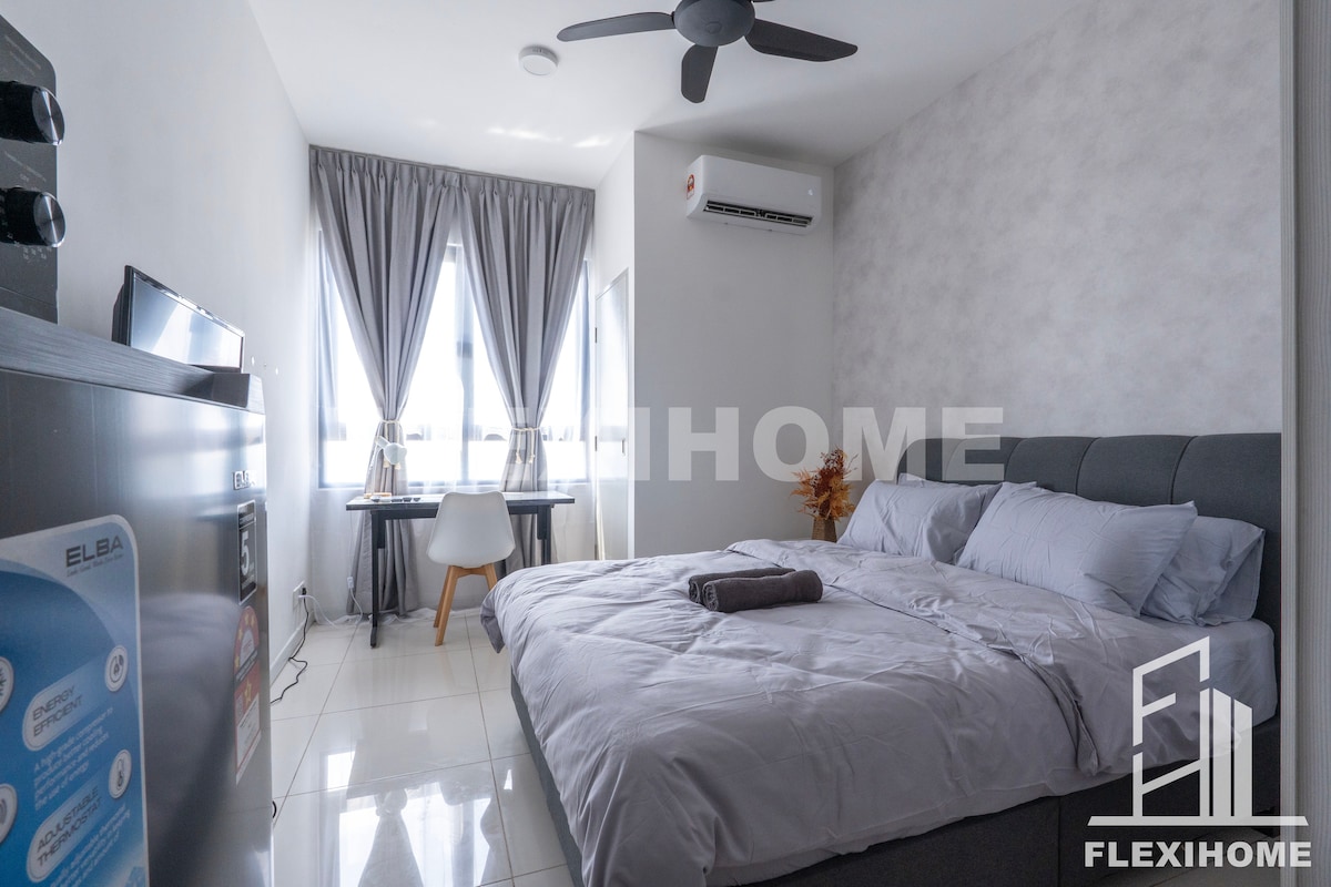 A cozy studio room is presented with a spacious bed covered in soft linens. A small desk adjacent to a window provides a workspace. Natural light filters in through the curtains, while a fan hangs from the ceiling, offering comfort in the bright, neutral-toned space.