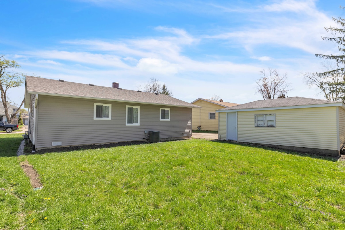 An exterior view of a single-story house is presented, highlighting the well-maintained yard with lush green grass. The home features light-colored siding and several windows, with a neighboring building visible to the right. Clear skies add a bright and open feel to the space.