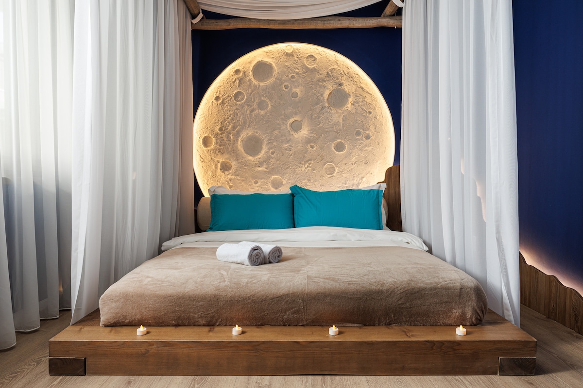 A serene bedroom is highlighted by a large moon artwork serving as a captivating backdrop. A spacious bed is dressed in soft linens with blue accent pillows. Decorative candles line the bed’s base, contributing to a calming environment. Sheer drapes softly frame the space.