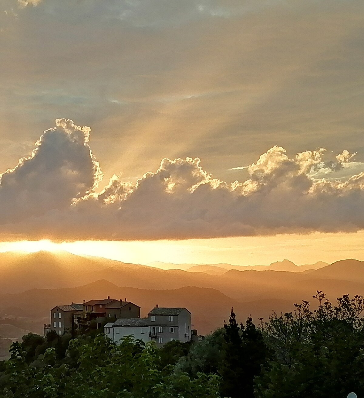A serene landscape captures a sunset view with soft rays of light breaking through clouds. Below, a cluster of houses is nestled amidst rolling hills, framed by distant mountains, creating a peaceful and scenic atmosphere.