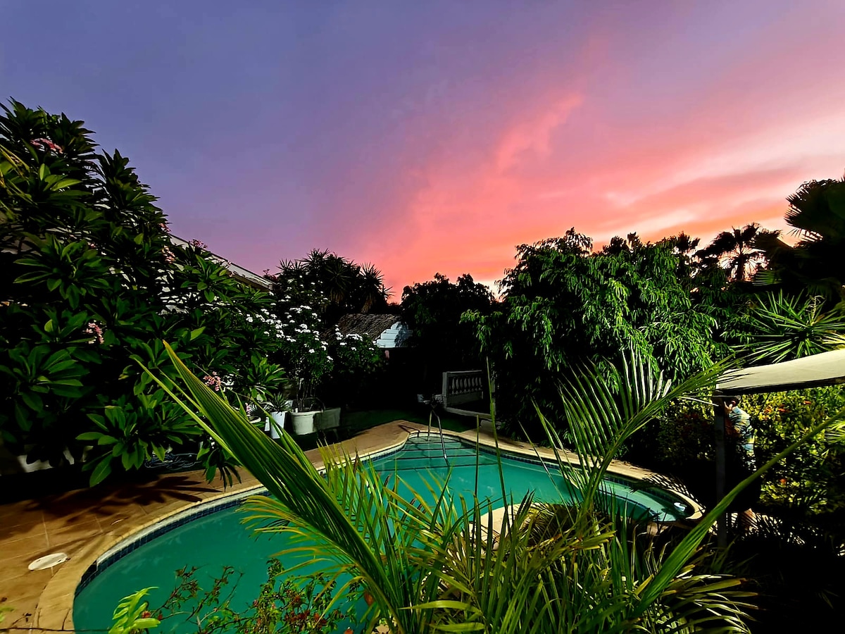A swimming pool is surrounded by lush greenery and vibrant flowers, reflecting the colors of a sunset sky. The patio area features seating options and is bordered by tall palm trees, creating a serene outdoor space.