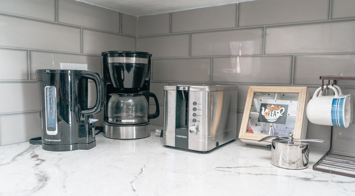 A modern kitchen countertop is showcased, featuring a kettle, coffee maker, and a silver toaster. A picture frame displaying a colorful image stands nearby, along with a small metal container. The glossy gray tiles in the background complement the marble surface.
