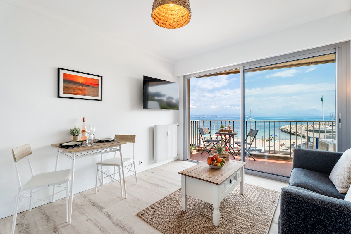 A bright living area features a modern dining table set for two and a comfortable sofa. Large glass doors open to a balcony with views of the sea. A simple coffee table sits atop a textured rug, complemented by light wood flooring and neutral walls.