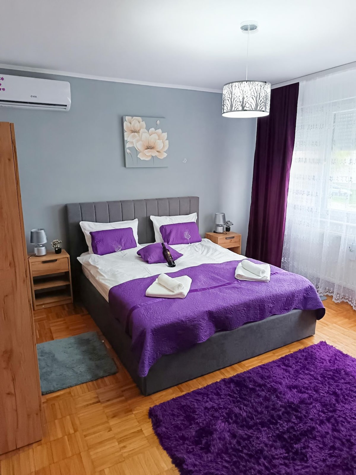 A serene bedroom is featured, with a spacious bed dressed in a light-colored blanket and accented by purple pillows. Nightstands with lamps are placed on either side. A window with sheer curtains allows natural light to illuminate the space, complemented by a decorative rug.