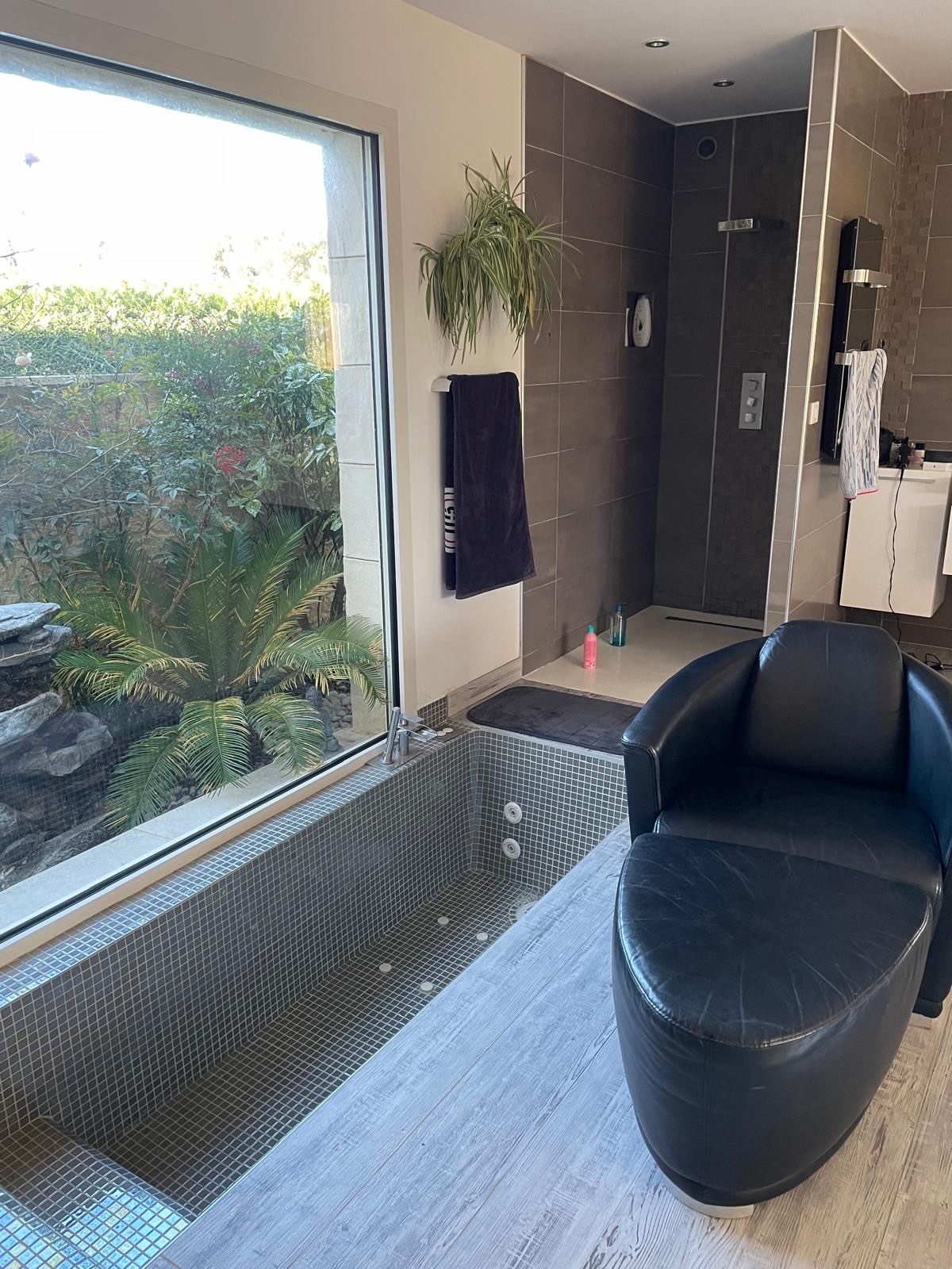 A modern bathroom features a sleek soaking tub with a view of a lush garden through expansive glass windows. A black lounge chair is positioned nearby, complementing the neutral tones of the tiled walls and flooring. A shower area is visible in the background.