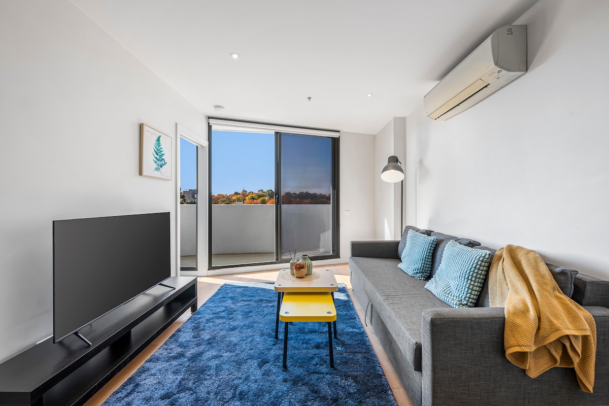 A bright living area features a cozy gray sofa adorned with textured blue cushions. A simple yellow coffee table sits in front of the sleek smart TV. Large glass doors open to a balcony, allowing natural light to fill the space and showcasing outdoor greenery.