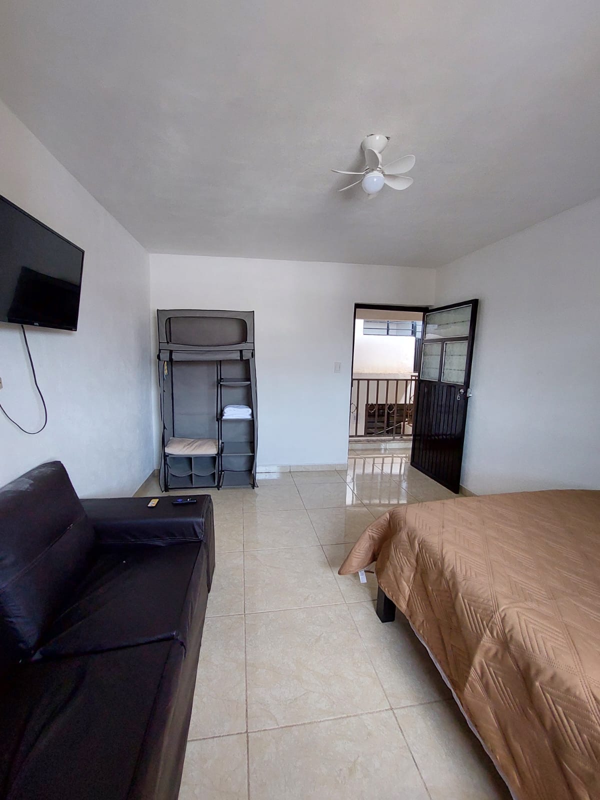 A room features a spacious bed with a neutral-colored cover and a cozy sofa. A wall-mounted television is visible, while a small shelving unit with towels is present in the corner. A doorway leads to an outdoor area, enhancing the room's functionality.