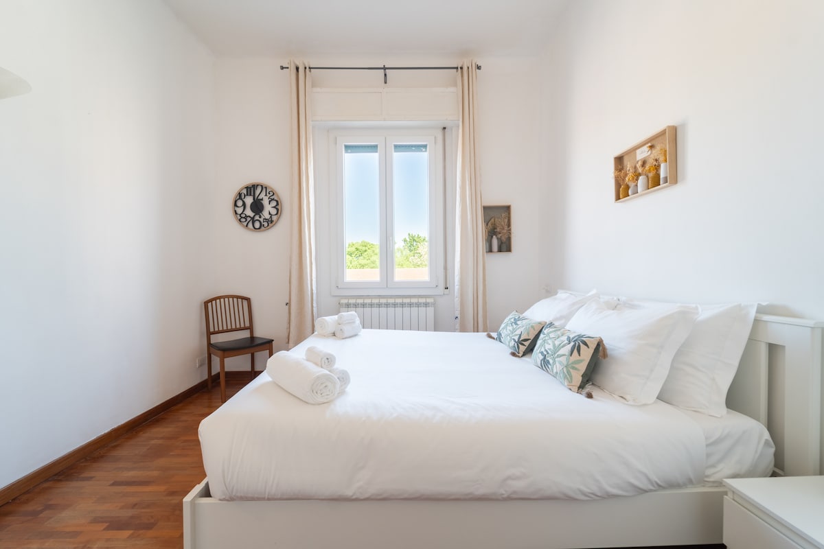 A spacious bedroom features a large bed with soft linens and decorative pillows. Natural light enters through two windows, complementing light-colored walls. A wooden chair and a wall clock enhance the simplicity, while towels are neatly arranged on the bed's edge.