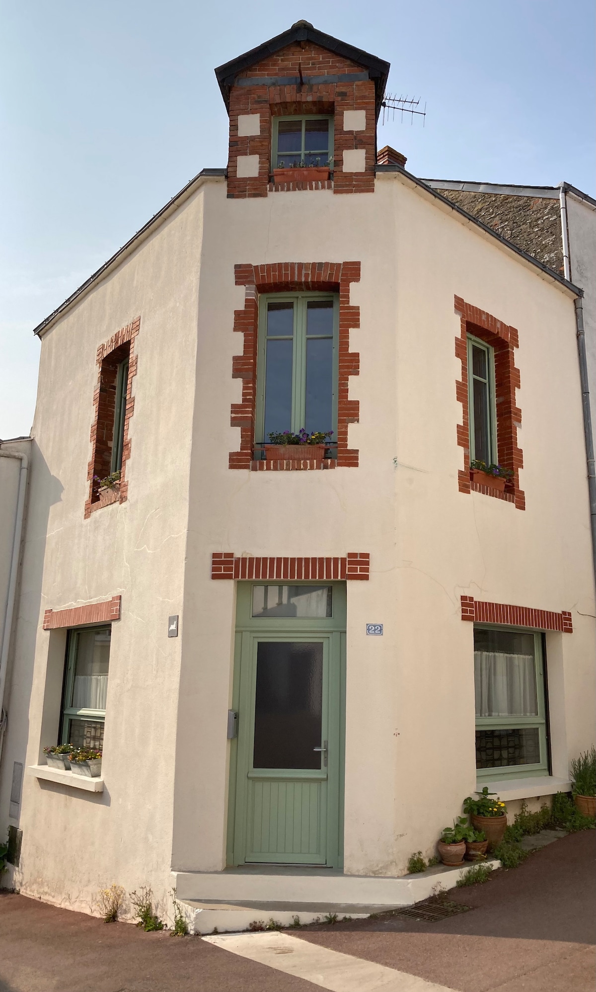 The charming exterior of the house is highlighted by its unique shape and distinct architectural details. Features include red brick accents around the windows and a green door. Flower pots adorn the window ledges, adding a touch of greenery to the facade.