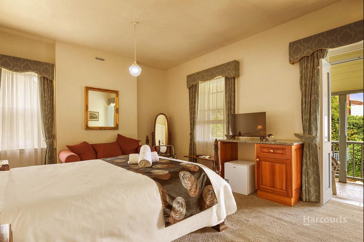 A spacious room is furnished with a large bed dressed in an elegant patterned bedspread. Natural light filters through sheer curtains, illuminating the area. A wooden dresser and a television are visible, along with a mirrored vanity and a cozy seating nook by the window.