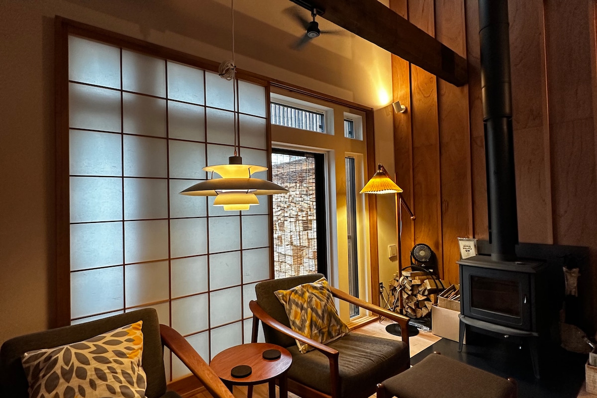 A cozy living area features two wooden chairs with patterned cushions, a small side table, and a wood-burning stove. Natural light filters through a shoji screen and a sliding glass door leads to a wood pile outside, creating a warm atmosphere.