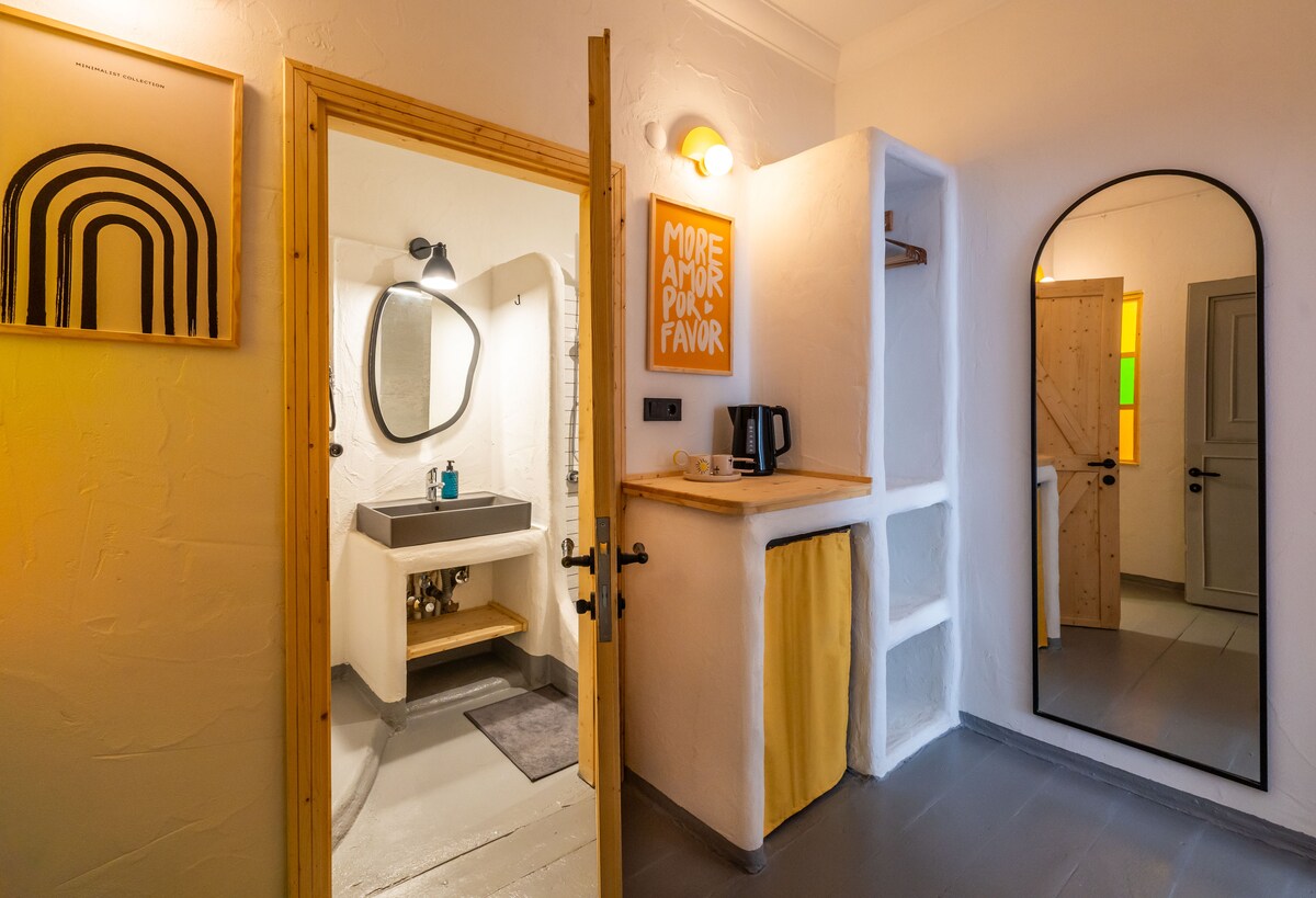 A bright room features a doorway leading to a bathroom with a modern sink and mirror. A small counter holds a kettle. An arch-shaped mirror reflects the space, enhancing the room's openness. Simple artwork adorns the wall, complementing a neutral color scheme.