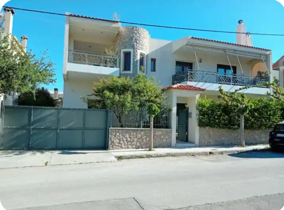 A modern two-story building is showcased with a stone facade and balconies adorned with railings. Lush greenery surrounds the property, creating a sense of privacy. A gated entrance is visible, along with a well-maintained path leading to the doorway.