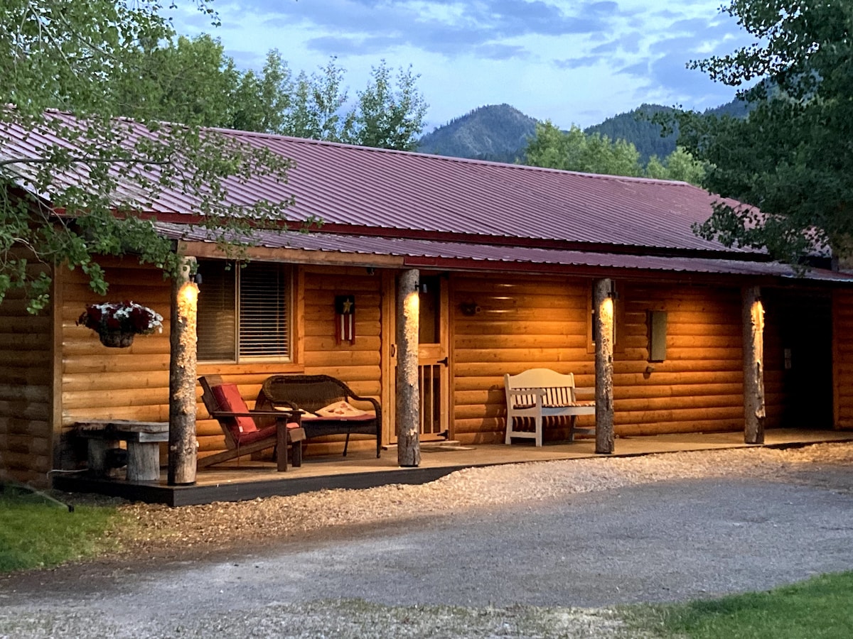 A rustic cabin exterior is showcased, featuring a red metal roof and wooden logs. A porch is furnished with a love seat and a bench, surrounded by trees and mountain views. Soft lighting highlights the entrance, creating an inviting ambiance.