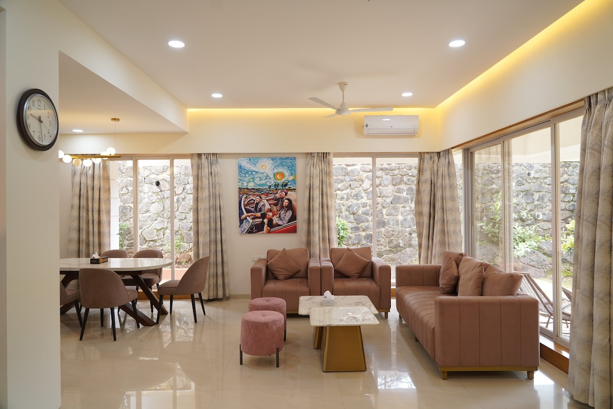 A spacious living area is highlighted by modern furniture, including a beige couch and pink accent chairs. Large sliding windows allow natural light to fill the room, providing views of the garden. A dining table is positioned nearby, accompanied by decorative wall art.