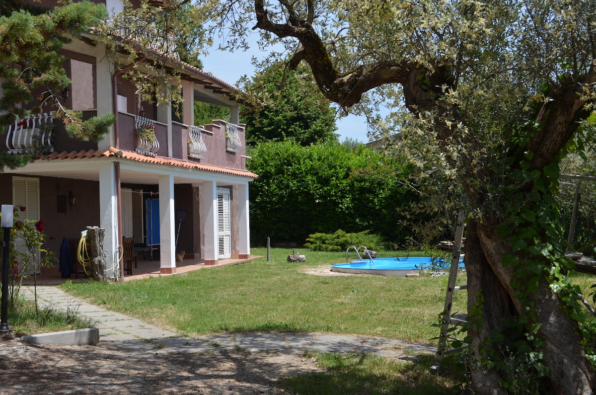 The image showcases a spacious garden surrounding a home with a patio and a pool. The pool is visible on the right, framed by green grass. The house features multiple balconies and large windows, providing a glimpse of the relaxing outdoor space.
