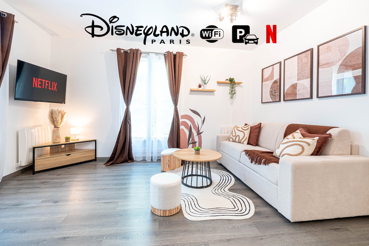 A cozy living area is presented with a neutral sofa adorned with patterned cushions. A wooden coffee table sits in the center, accompanied by two small stools. A wall-mounted TV displays the Netflix logo. Light filters through sheer curtains, enhancing the space's brightness.
