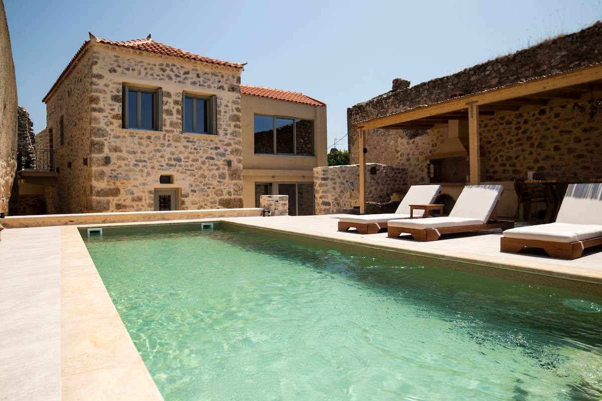 An infinity pool is featured, merging with the landscape, surrounded by wooden sunbeds with comfortable mattresses. The rustic stone villa provides a striking backdrop, while large glass windows reflect the outdoor area, allowing natural light to illuminate the space. A shaded barbecue area is visible to one side.