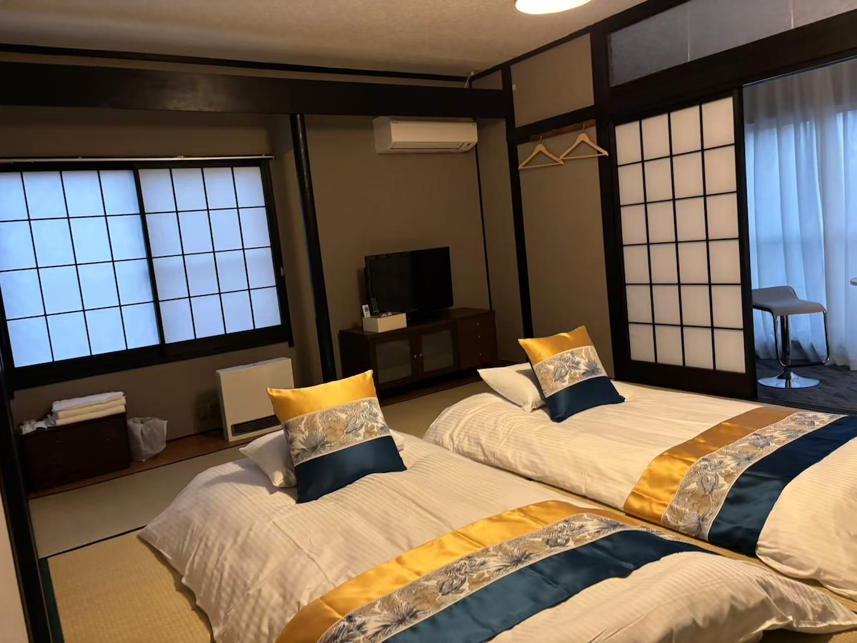 A tranquil room features two spacious beds adorned with elegant bedding in soft tones of white and gold. Natural light filters through shoji screens, creating a serene atmosphere. A small television is positioned on a wooden console, along with a hanger for easy storage.
