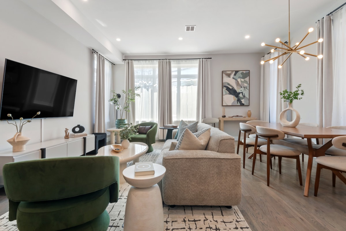 The living area features a modern design with a spacious seating arrangement, including a light gray sofa and green accent chairs. A large flat-screen TV is mounted on the wall, while natural light floods the room through tall windows. A dining table for six is positioned beside the seating area, all complemented by tasteful décor.