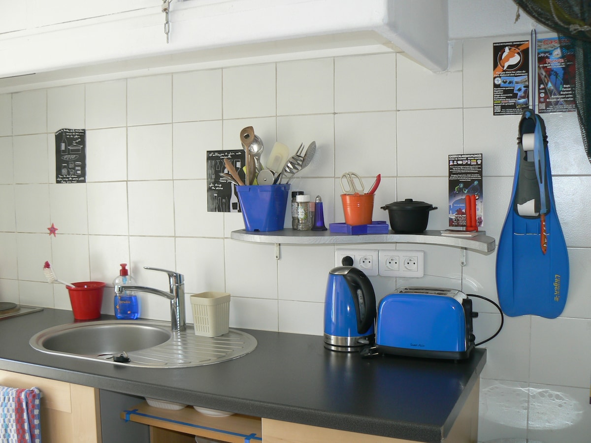 The kitchen features a modern countertop with a silver sink and an array of cooking utensils displayed in colorful containers. An electric kettle in blue and a black cooking pot are positioned for easy access. Small appliances are neatly arranged, contributing to a functional workspace.