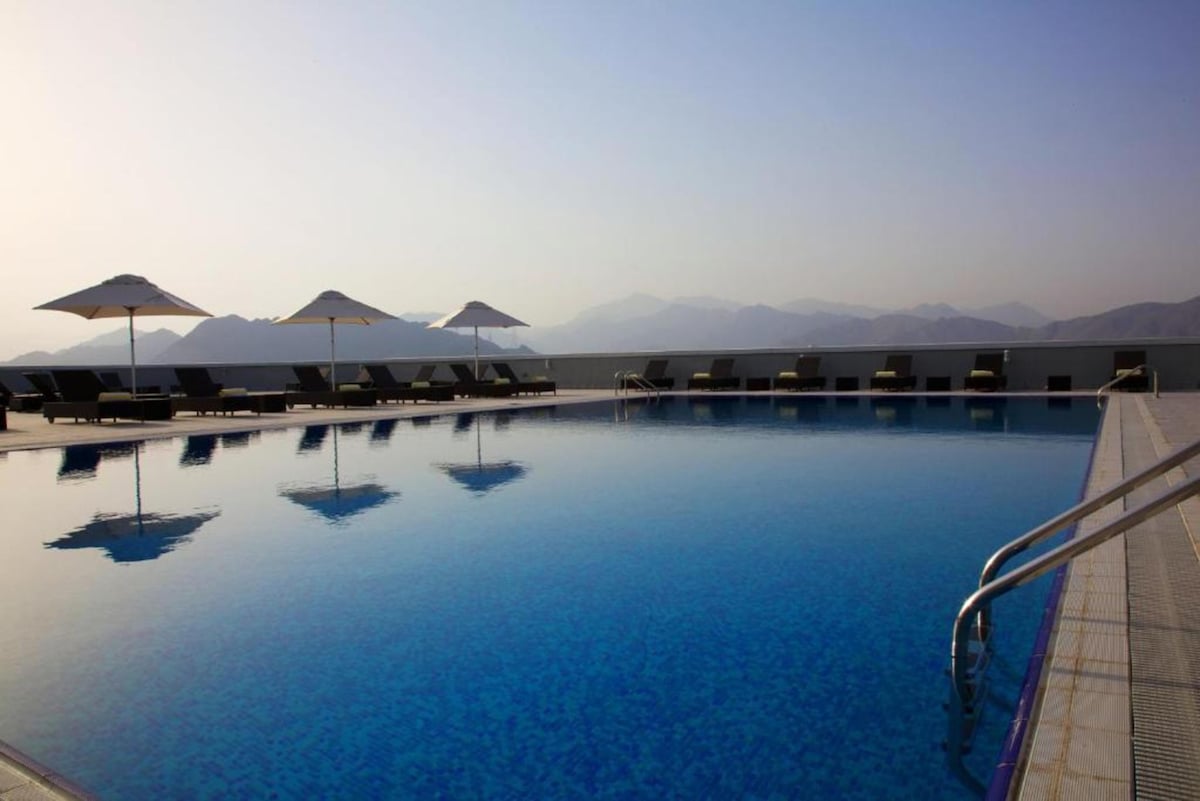 A rooftop pool is surrounded by lounge chairs and umbrellas, reflecting the serene mountains in the water. The clear sky above casts soft light on the pool area, creating a tranquil setting ideal for relaxation.