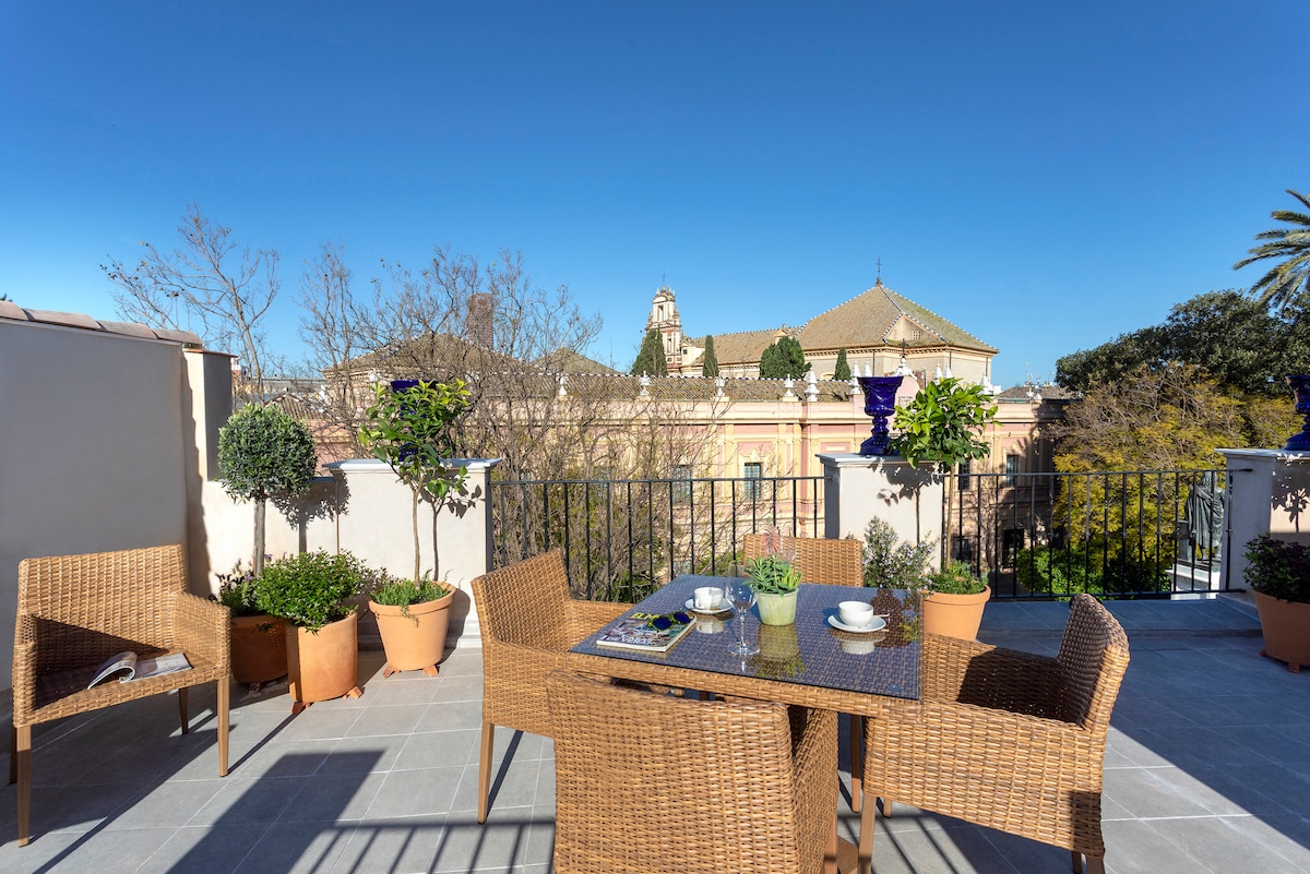 An outdoor terrace is visible, featuring a table surrounded by four wicker chairs. Potted plants add greenery to the space, which overlooks nearby architectural structures under a clear blue sky.