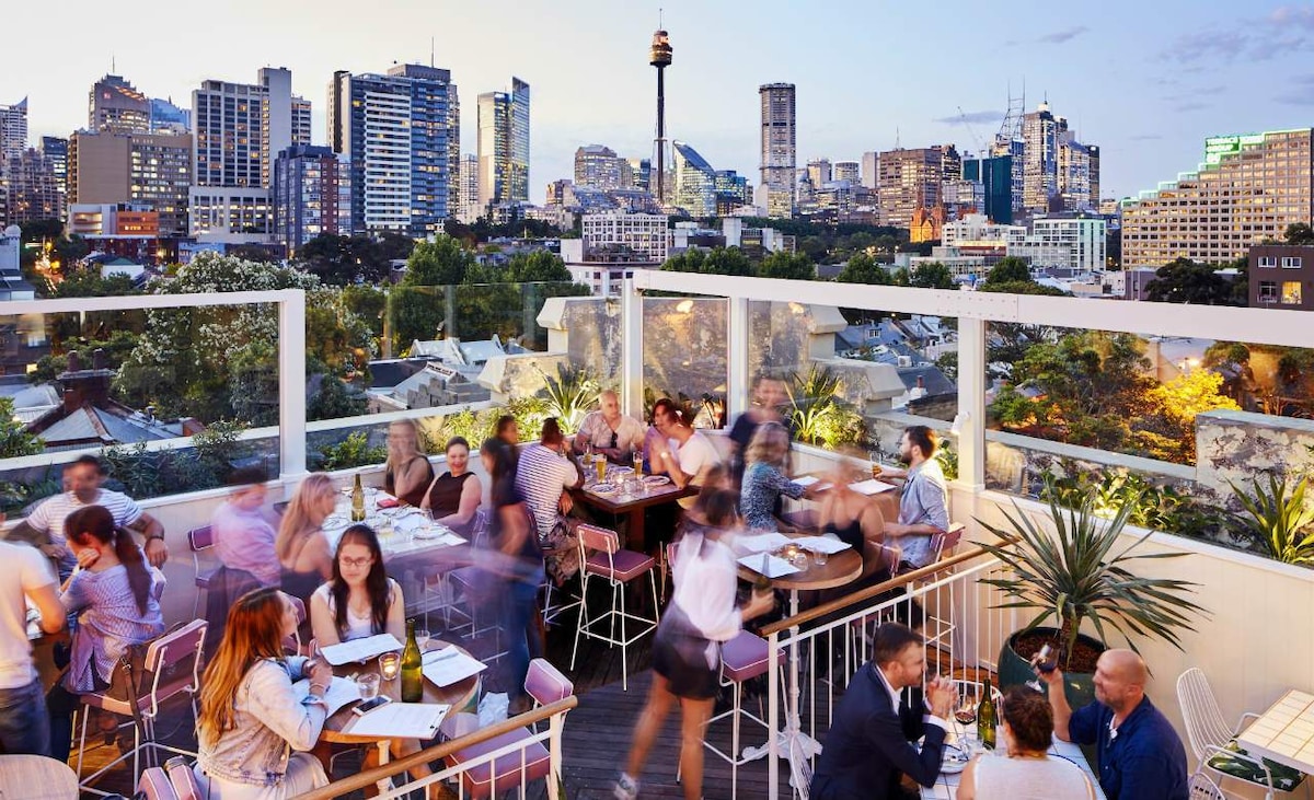 A lively rooftop dining space is showcased, with tables filled with diners enjoying the vibrant atmosphere. The skyline of Sydney is visible in the background, highlighting the urban landscape, with the iconic tower prominently displayed against a dusky sky.