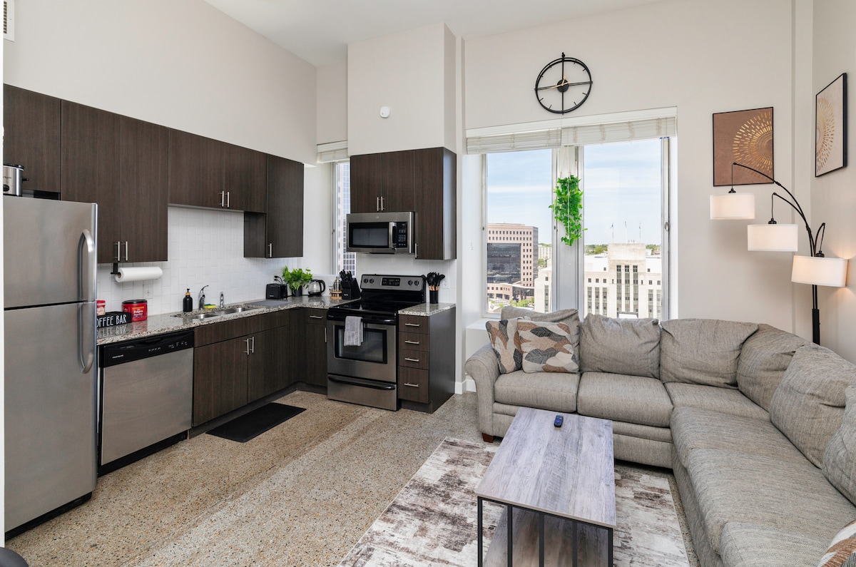 The spacious kitchen features modern appliances, including a refrigerator and stove, with a sleek design in dark cabinetry. A large sectional couch is situated in the living area, accompanied by natural light from windows that offer a city view. A wall clock and minimalist décor enhance the contemporary look.