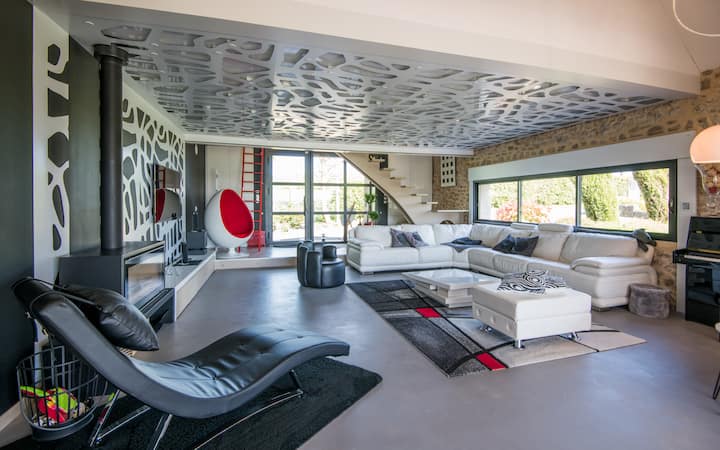 Salon design et lumineux avec vue sur le jardin.
Le salon, à l'identité architecturale originale offre une atmosphère moderne et raffinée. La décoration noir béton et blanc, ponctuée de touches de rouge, ajoute de la chaleur à l’ensemble. 