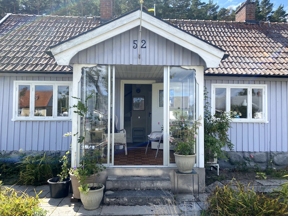 The entrance of a charming house features a welcoming porch with glass doors and potted plants. The purple exterior is complemented by a tiled roof. Two chairs are positioned on the porch, inviting guests to enjoy the surroundings.