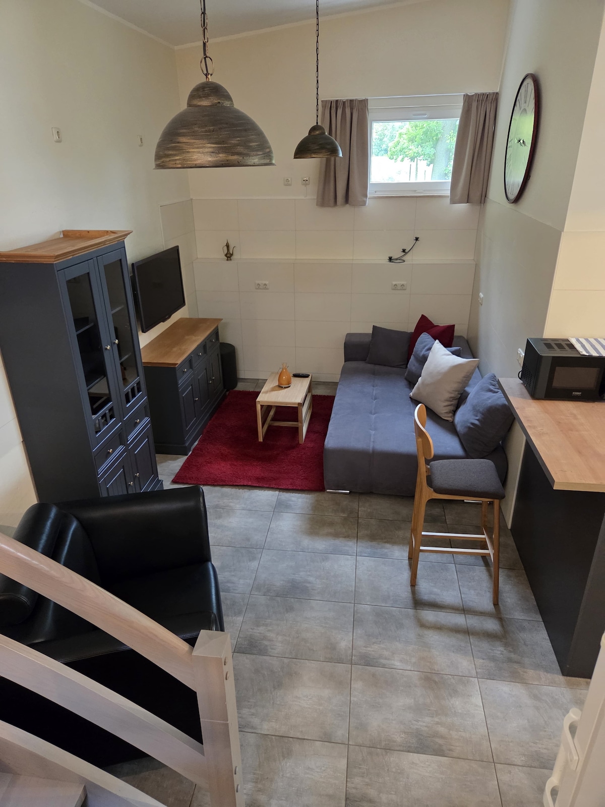 A cozy living area features a gray sofa and an armchair, accompanied by a low wooden coffee table and a red area rug. A light wooden cabinet holds a flat-screen TV. Natural light enters through a window adorned with light curtains, enhancing the space.
