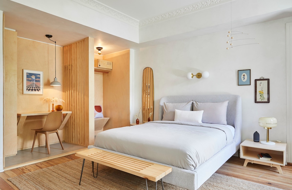 A spacious room features a light-colored queen bed with a soft duvet and decorative pillows. A minimalistic desk and chair set are situated nearby, complemented by a soft light fixture. Natural wood accents enhance the design, and a variety of art pieces are displayed on the walls.