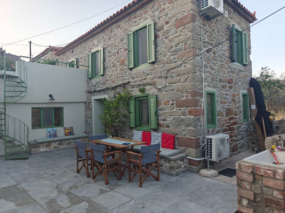 The traditional stone house features a spacious outdoor area with a large stone patio. An outdoor dining table surrounded by wooden chairs is visible, along with green shutters on the windows, and a spiral iron staircase leading to the upper level.