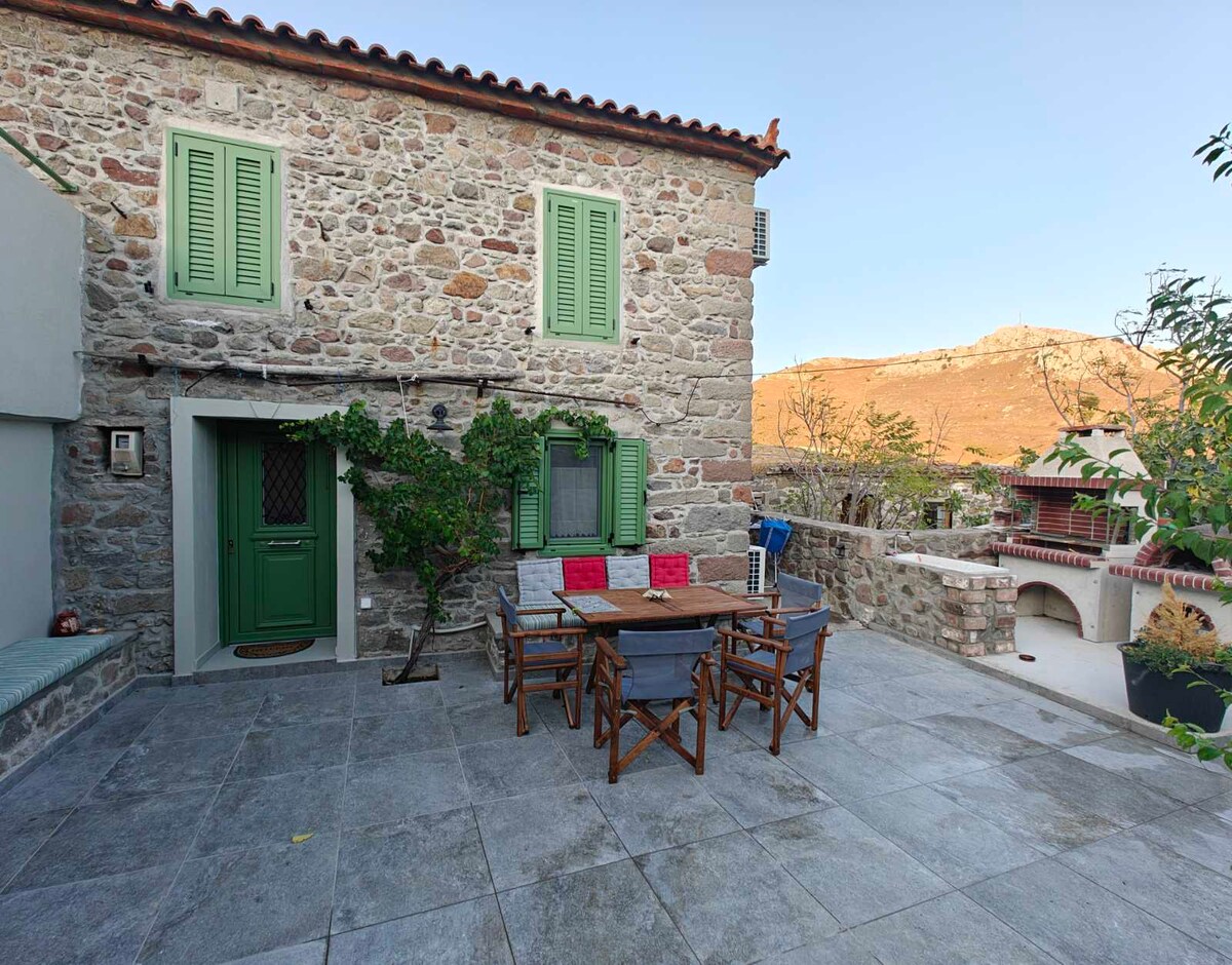 A traditional stone house features green shutters and a welcoming entrance. Outdoor seating is provided around a wooden table, accompanied by four folding chairs. The spacious stone-paved courtyard offers views of the surrounding landscape and is adorned with greenery.