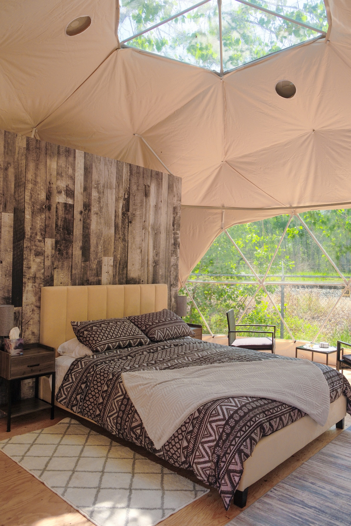 A cozy sleeping area features a spacious bed adorned with patterned bedding. A wooden accent wall creates a natural feel, while large windows allow ample natural light. A small bedside table is positioned beside the bed, enhancing the functional layout of the room.