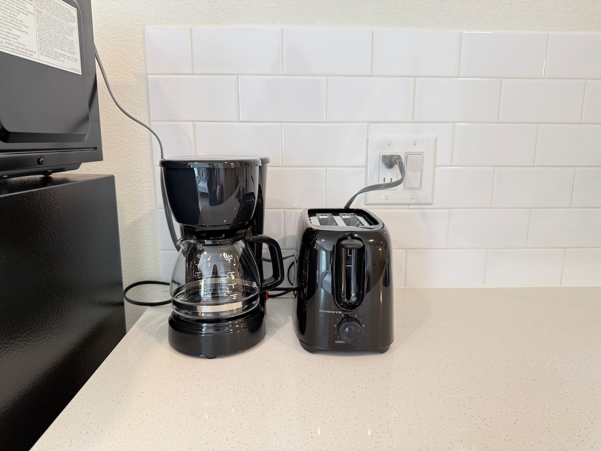 A modern kitchen countertop features a black coffee maker and toaster, positioned against a backdrop of white subway tiles. The countertop is smooth and light-colored, providing a clean, functional space for meal preparation.