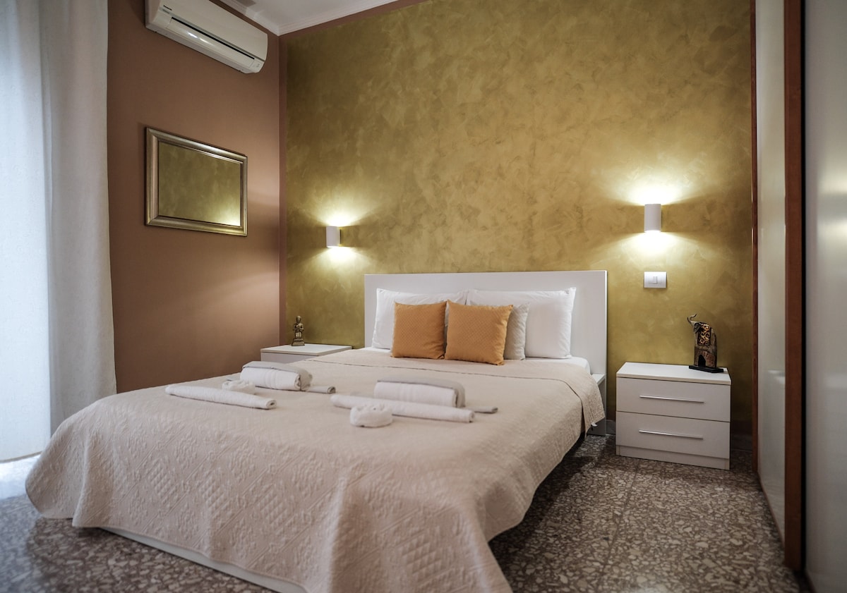 A bright sleeping area features a spacious bed adorned with light bedding and decorative cushions. Flanking the bed, two bedside tables provide additional storage. Soft lighting from wall sconces complements the warm gold-hued accent wall, enhancing the calming atmosphere.