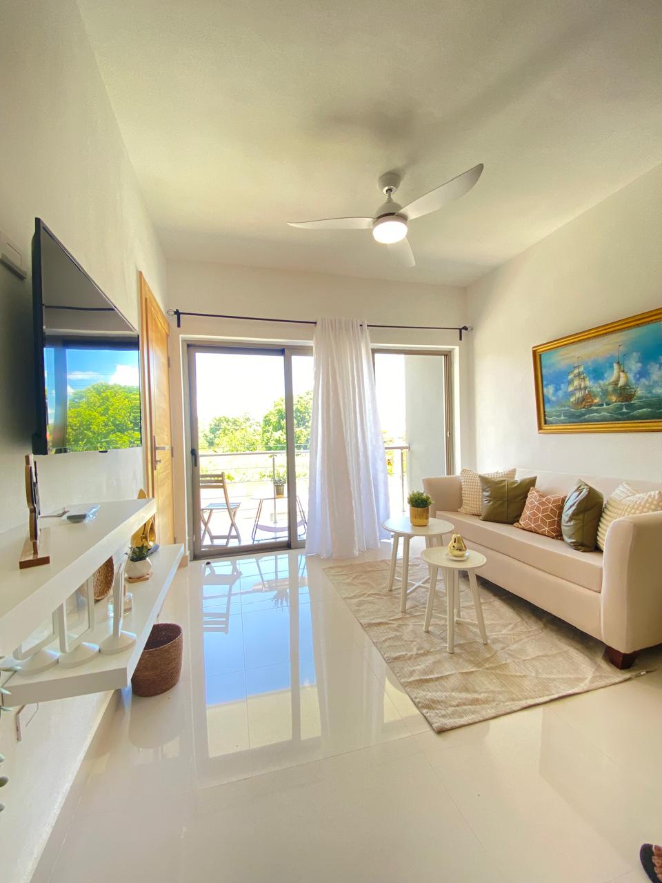 A bright living area features a plush sofa complemented by decorative pillows. A coffee table sits in front, and a large window opens to a balcony showcasing natural views. Light reflects off the glossy floor, enhancing the spacious ambiance.