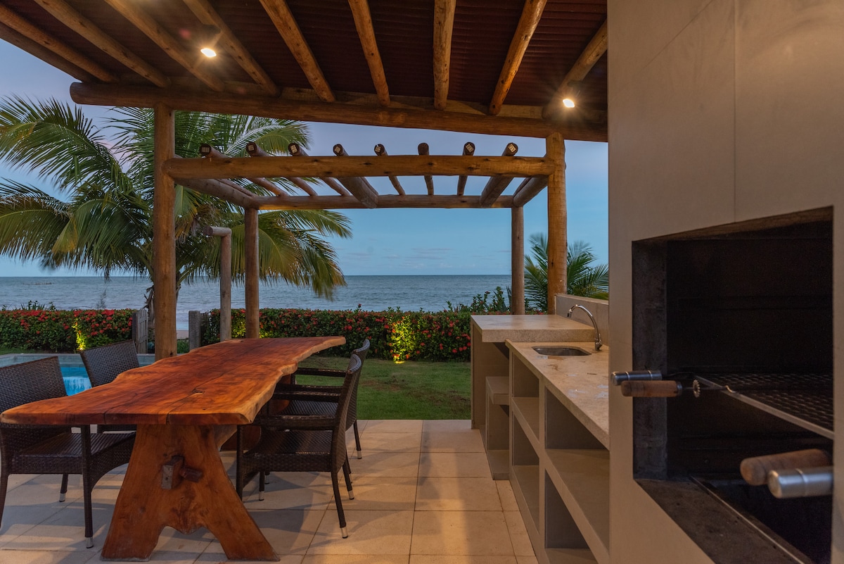 An outdoor barbecue area features a wooden dining table surrounded by chairs, with a built-in grill and sink nearby. The space is framed by tropical plants and offers a view of the calm ocean, creating a relaxing setting for gatherings.
