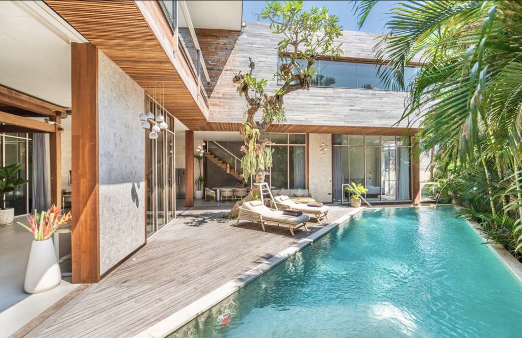 An outdoor area showcases a sleek pool bordered by wooden decking and tropical plants. Sun loungers are positioned beside the pool, inviting relaxation. Large glass panels allow natural light to enter, seamlessly connecting the indoor and outdoor spaces within a contemporary architectural design.