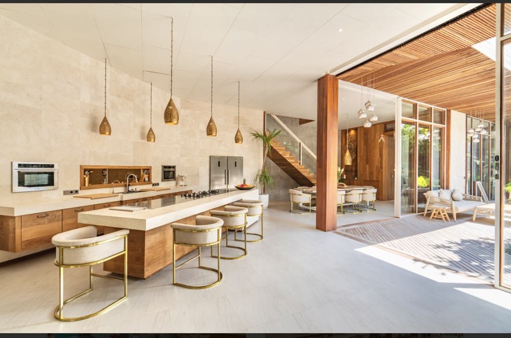 A modern kitchen is highlighted by sleek cabinetry and a central island with gold-accented bar stools. Pendant lights hang above the island, and large glass doors allow natural light to fill the space. A dining area is visible in the background, seamlessly integrated into the open layout.