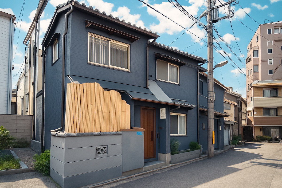 The exterior of a two-story, modern house features a dark blue facade with wooden accents. A bamboo fence lines the entrance, adding a touch of traditional design. Surrounding utilities and neighboring buildings are visible against a clear blue sky.