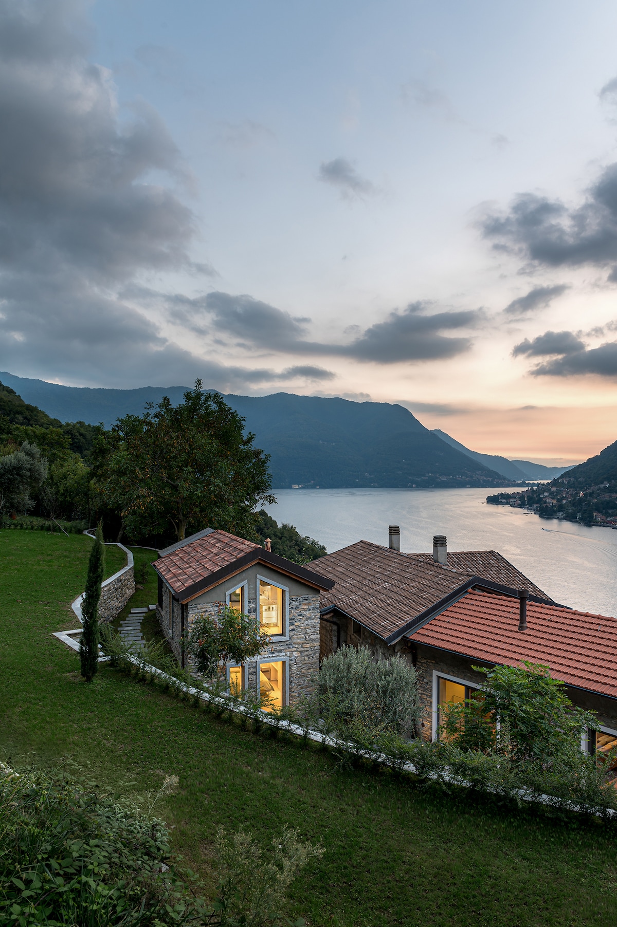 A charming stone house is nestled on a hillside, surrounded by lush greenery. Large windows reflect the warm glow from the interior, while breathtaking views of Lake Como and distant mountains are visible in the background, bathed in soft twilight hues.