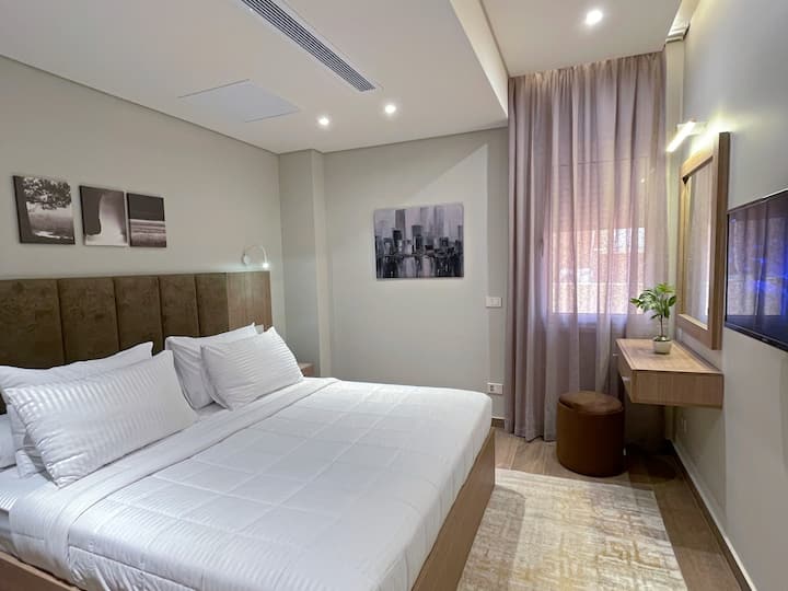 غرفة نوم مريحة تضم سرير أنيق، فرش عالي الجودة، أثاث متناسق، ديكور راقٍ، إضاءة ناعمة، بياضات نظيفة، ستائر فاخرة، سجاد أرضيات أنيق، وشاشة مسطحة لراحة واستمتاع الضيوف